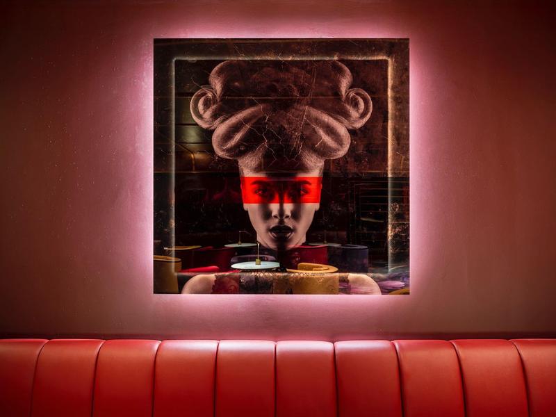 Stylish wall art of an unusual face above a red seating area in dim lighting.