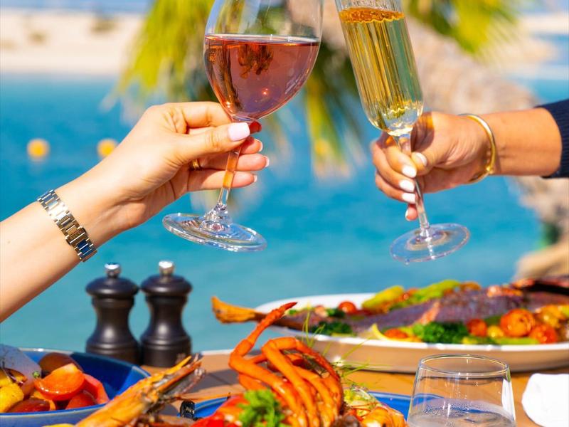 Two people clinking wine and champagne glasses at a table set with seafood and a sea view.
