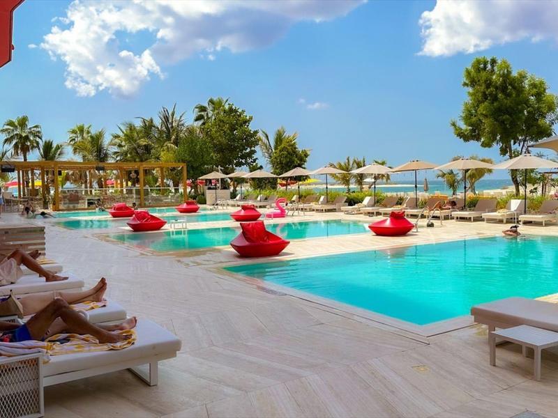 Modern outdoor pool with red loungers and palm trees at hotel under sunny sky.