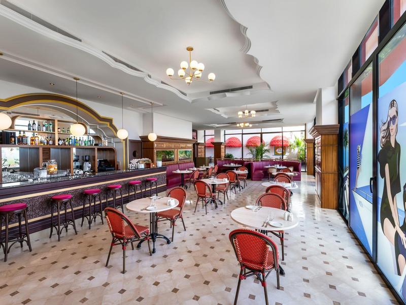 Bright hotel bar with red chairs, tables, and a long bar with stools.