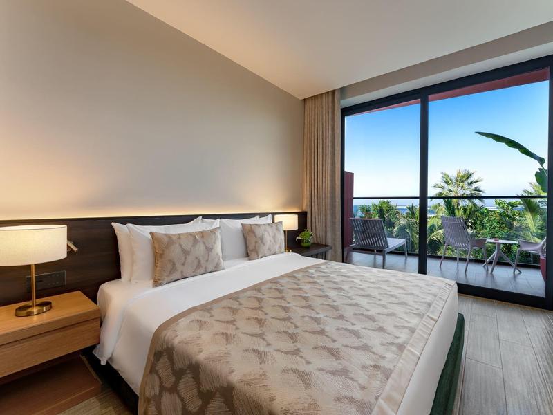 Modern hotel room with double bed, bedside lamps, and balcony overlooking palm trees.