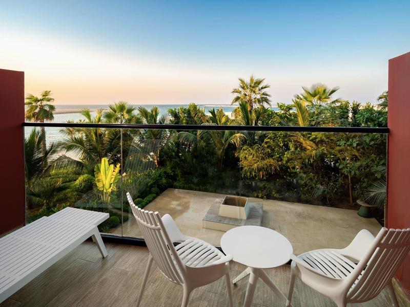 Balcony with white chairs and table overlooking palm trees and the sea at sunset.