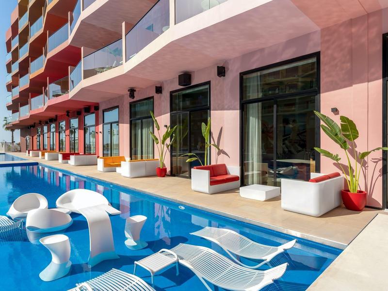 Modern pool area with white loungers and seating in front of a pink multi-story hotel.