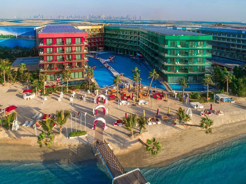 Colorful resort with pool by sandy beach and blue lagoon, skyline in the background.