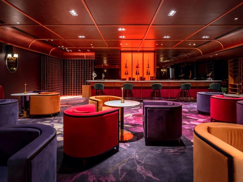 Elegant hotel bar with red and purple armchairs, reddish lighting, and a round carpet.