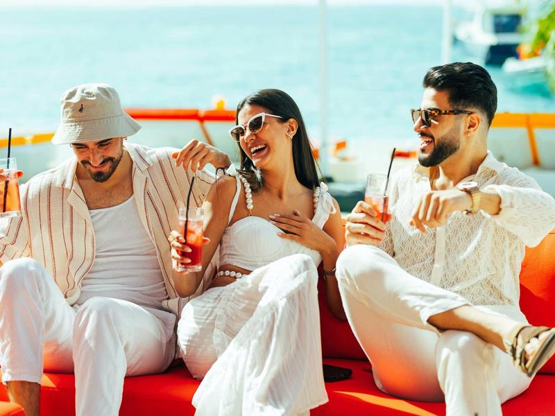 Three friends relax on red seats by the sea with summer drinks.