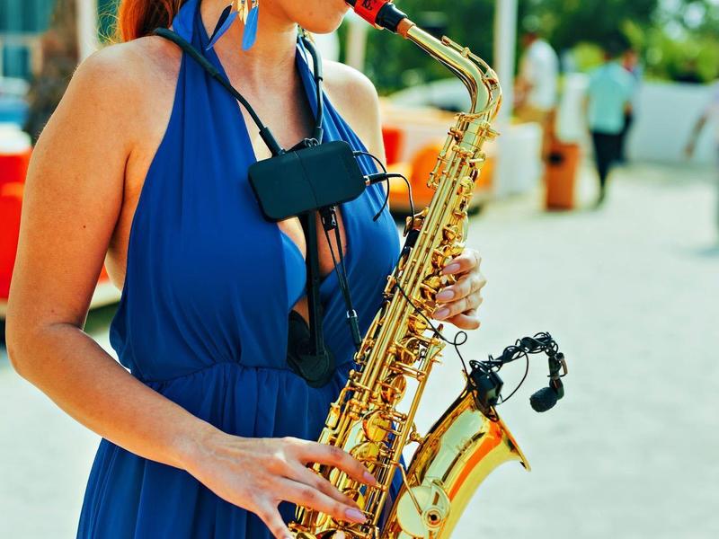 Woman in blue dress playing saxophone outdoors on a sunny day.