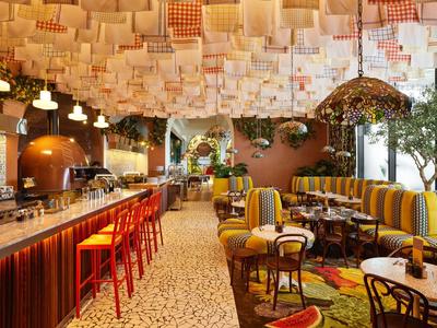 Buntes Restaurant mit gepolsterten Sitzbänken, roten Barhockern, Holztischen und strukturiertem Deckenmuster.