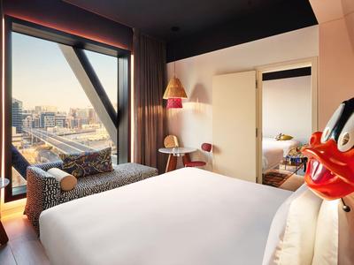 Modernes Hotelzimmer mit großem Fenster, weißem Bett, roter Lampe und urbaner Stadtansicht.