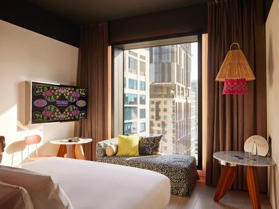 Modernes Hotelzimmer mit großem Bett, Fernseher, Fenstersitz und Blick auf Hochhäuser.