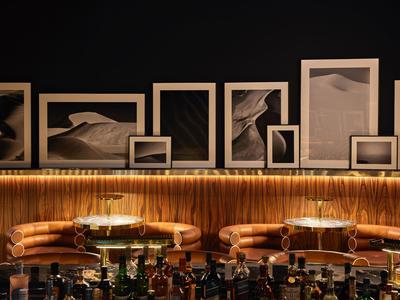 Moderne Bar mit braunen Ledersesseln, beleuchtetem Holztresen und Schwarz-Weiß-Wüstenfotos an Wand.
