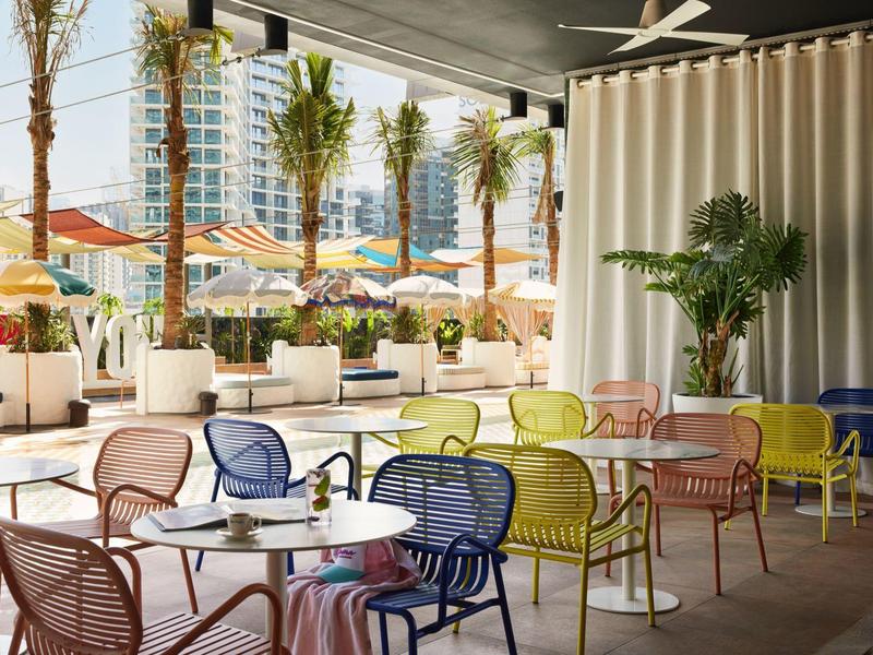 Colorful chairs and tables under a café roof with views of palm trees and sun umbrellas.