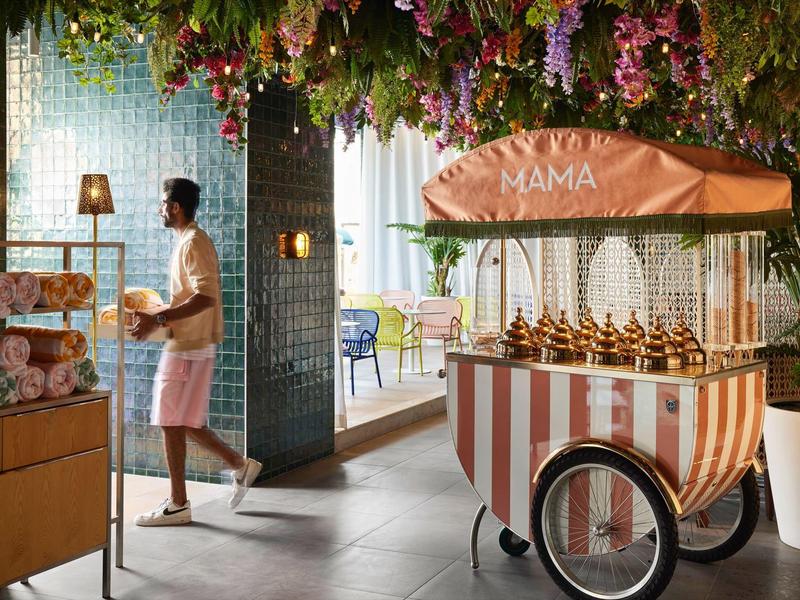 A man in a bathrobe takes fresh bread rolls from a small cart in a hotel area with flowers and towels.