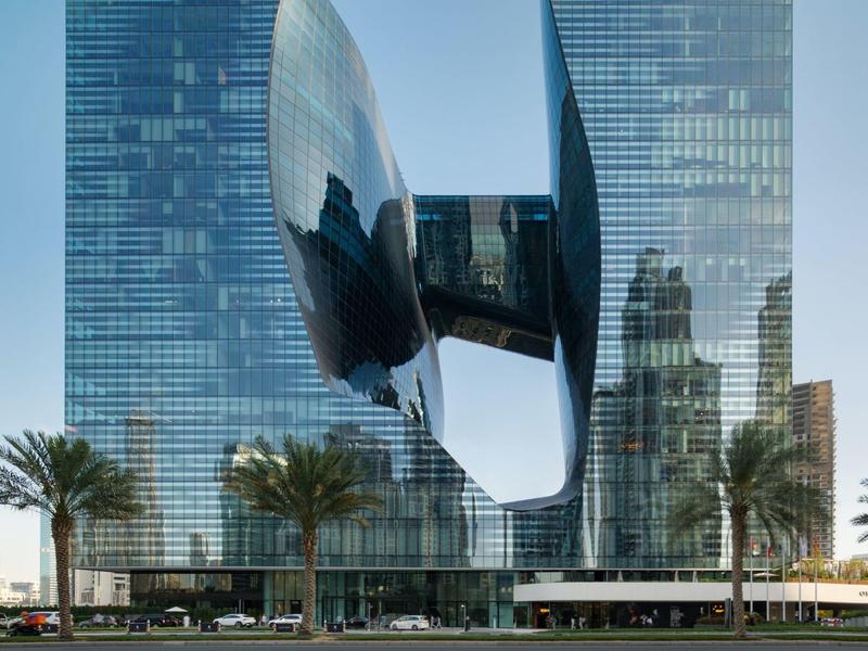 Modern glass twin towers connected by a curved skybridge, palm trees in front on a clear day.