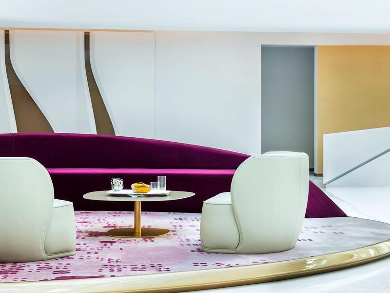 Modern lounge area with white chairs, circular tables, and a purple curved sofa in a bright room.