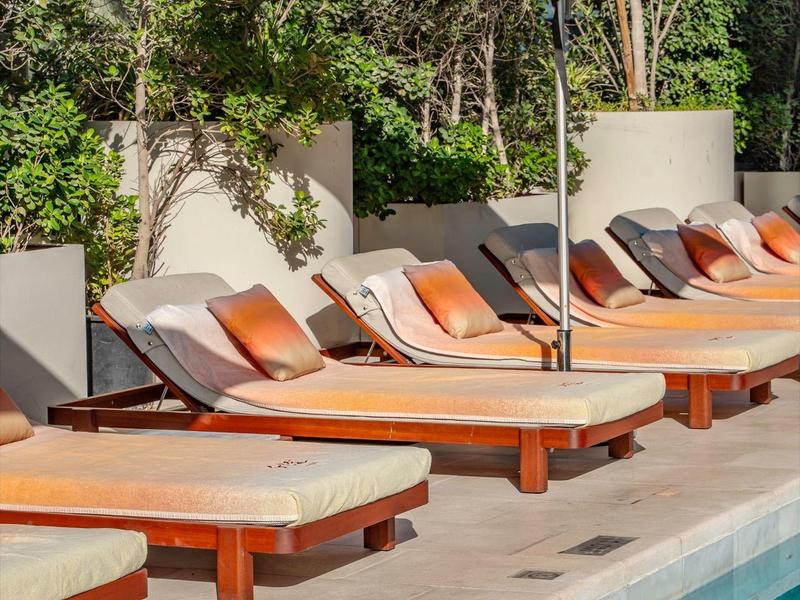 Poolside lounge chairs with cushions under large umbrellas by a clear pool.