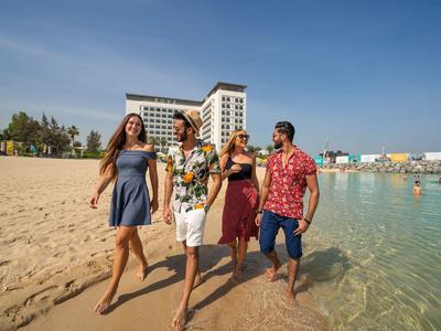 Four people walk barefoot along a sandy beach by clear blue water with a hotel in the background.