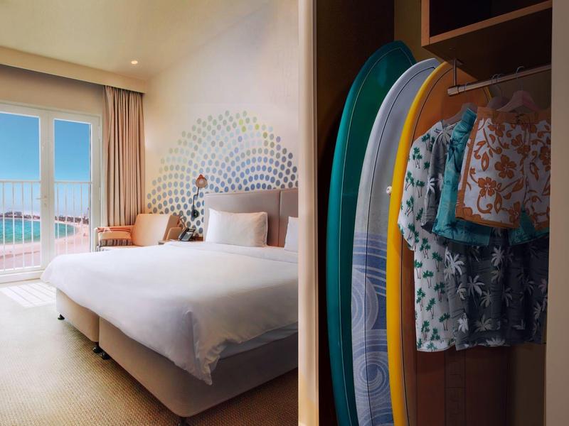 Bright hotel room with large bed, colorful surfboards, and balcony with ocean view.