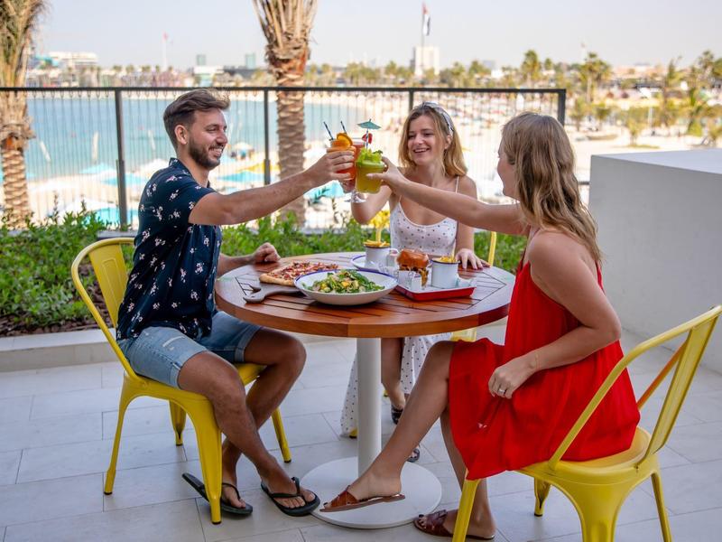 Three people sit outdoors on yellow chairs around a table with food, toasting drinks by a waterfront.
