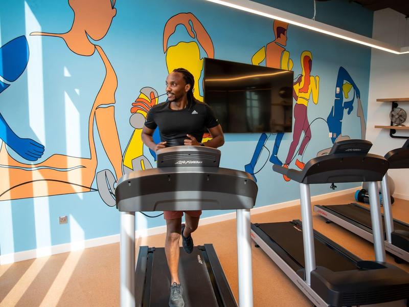 Man running on treadmill in gym with colorful wall mural and TV.