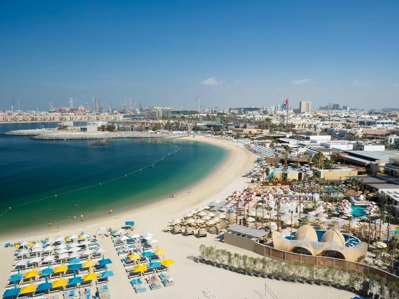 A beach resort with colorful umbrellas, sunbeds, and a city in the background under a clear sky.