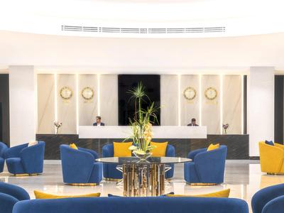 Světlý hotelový salonek s modrými a žlutými křesly kolem kulatých stolů s rostlinami.