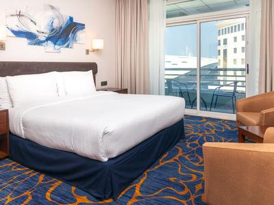 Moderně zařízený hotelový pokoj s velkou postelí a balkonem s výhledem na městské budovy.