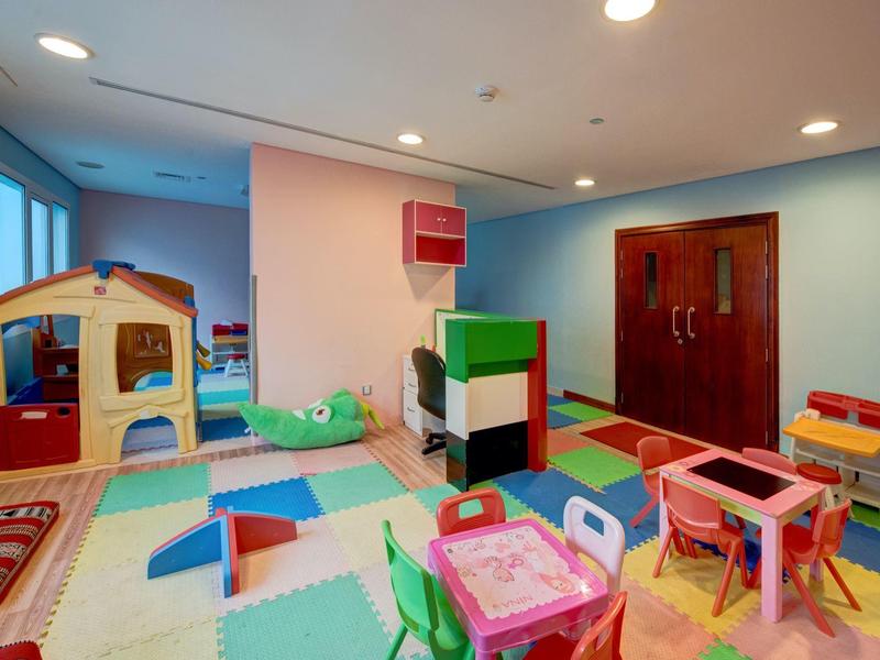 Barevná dětská herna s domečky na hraní, stoly, židlemi a měkkými podlahovými podložkami.
