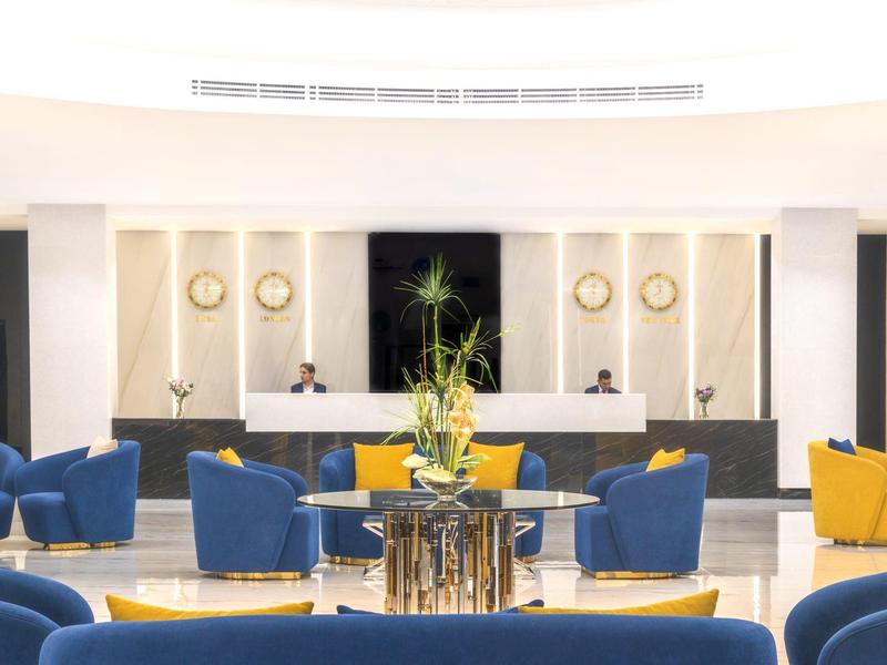 Světlý hotelový salonek s modrými a žlutými křesly kolem kulatých stolů s rostlinami.