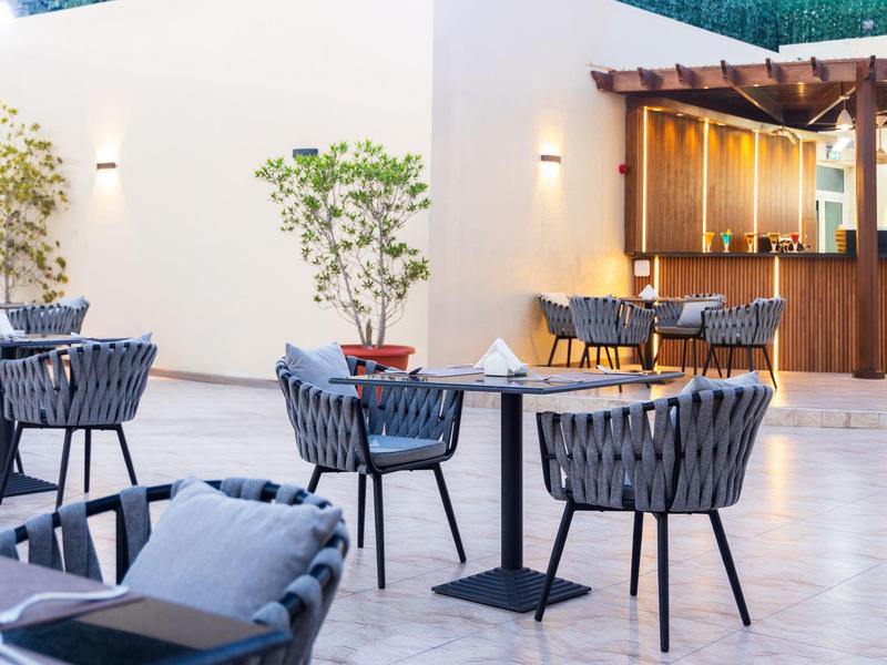 Moderní venkovní prostor hotelu se stoly, židlemi a barem pod pergolou.