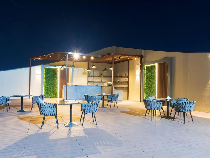 Moderní venkovní bar se osvětlenými vertikálními zelenými stěnami a modrými židlemi pod jasnou noční oblohou.