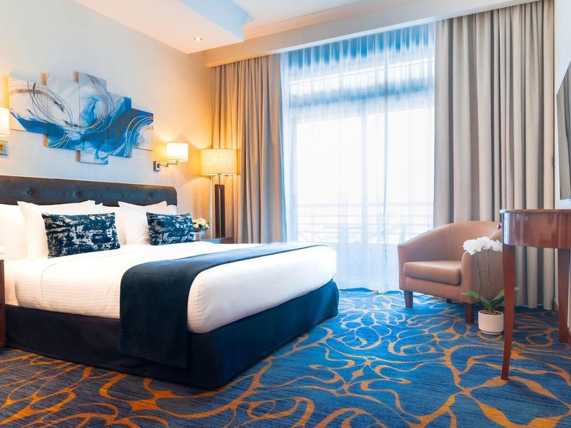 Moderní hotelový pokoj s manželskou postelí, křesly, kobercem a velkým oknem s záclonami.