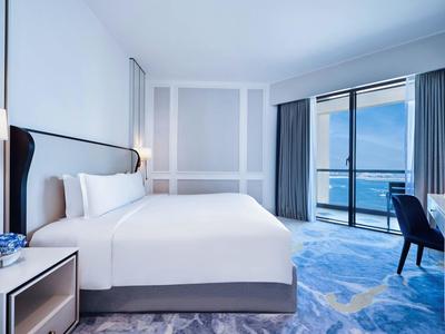 Habitación de hotel moderna con cama grande, mesita, silla y vista al mar a través de grandes ventanas.