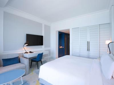 Habitación de hotel moderna con cama grande, escritorio, sofá y armario blanco, iluminación natural.