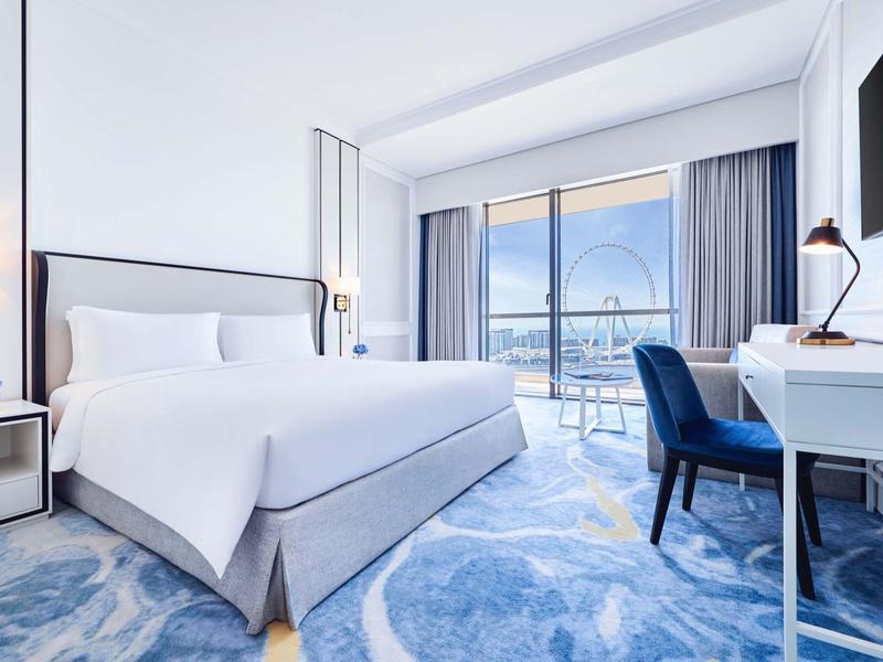 Habitación de hotel luminosa con cama grande, escritorio, silla azul y balcón con vista al mar.