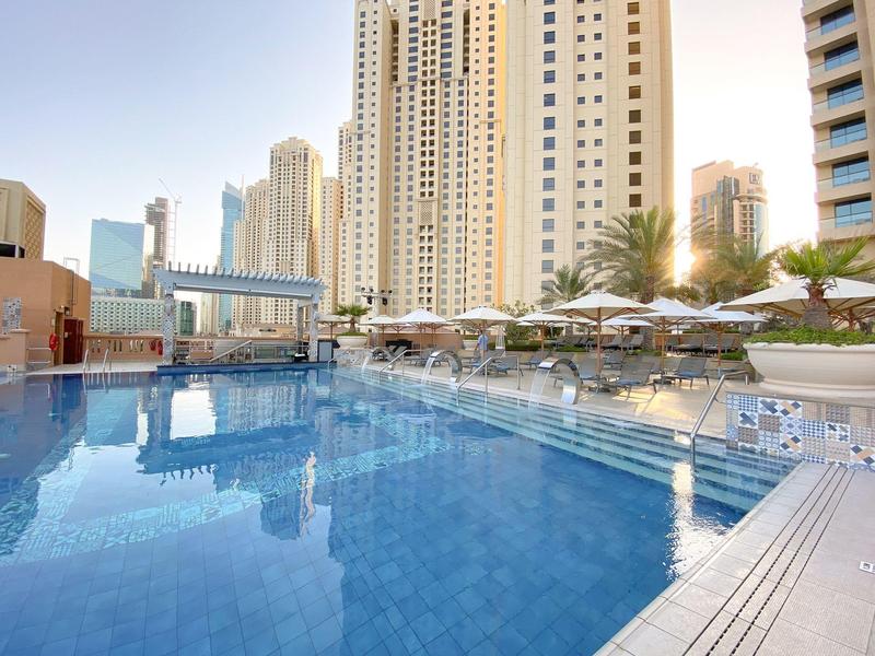 Gran piscina al aire libre con sombrillas y rascacielos al fondo bajo cielo despejado.