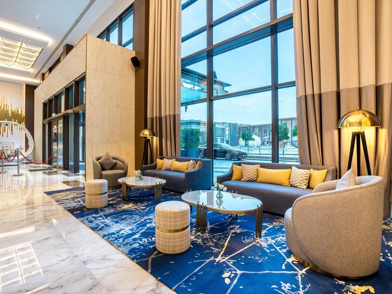 Área elegante del vestíbulo del hotel con grandes ventanas, muebles modernos y alfombra azul.