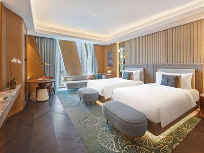 Modern hotel room with two beds, armchairs, carpet, and large windows with curtains.