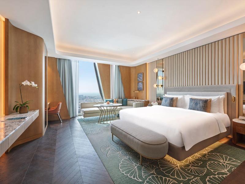 Bright hotel room with large bed, sofa, rug, and window with an outside view.