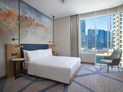 Jasny pokój hotelowy z dużym łóżkiem, krzesłem przy oknie i widokiem na miasto.