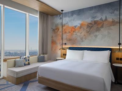 Modernes Schlafzimmer mit großem Fenster, gemütlichem Bett, Wandkunst und Sitzbereich mit Stadtblick.