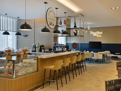 Moderner Cafébereich mit langer Holztheke, Barhockern, Hängelampen und Sitzgelegenheiten.