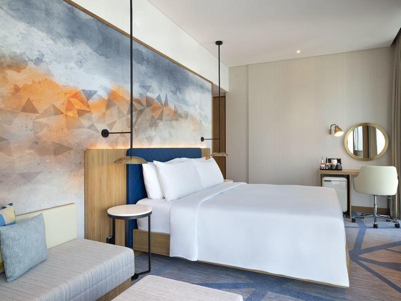 Modernes Schlafzimmer mit großem Bett, abstrakter Wandkunst, Sofa und Arbeitsplatz mit Spiegel.