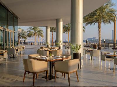 Terrasse eines Hotels mit Tischen und Stühlen, Palmen und Blick auf das Wasser und die Stadt im Hintergrund.