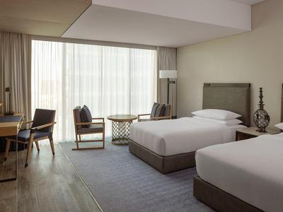 Moderní hotelový pokoj se dvěma postelemi, posezením a velkým oknem se záclonami.