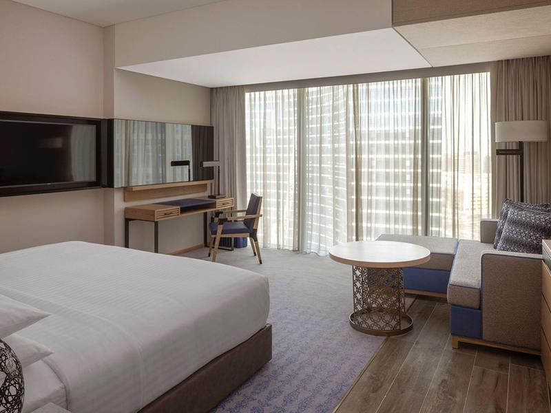 Moderní hotelový pokoj s velkou postelí, pohovkou, psacím stolem a okny od podlahy ke stropu.
