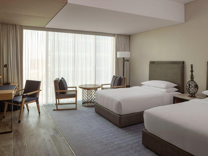 Moderní hotelový pokoj se dvěma postelemi, posezením a velkým oknem se záclonami.