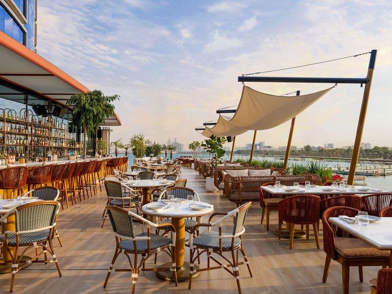 Venkovní restaurace s stoly, židlemi a slunečními plachtami u vody.