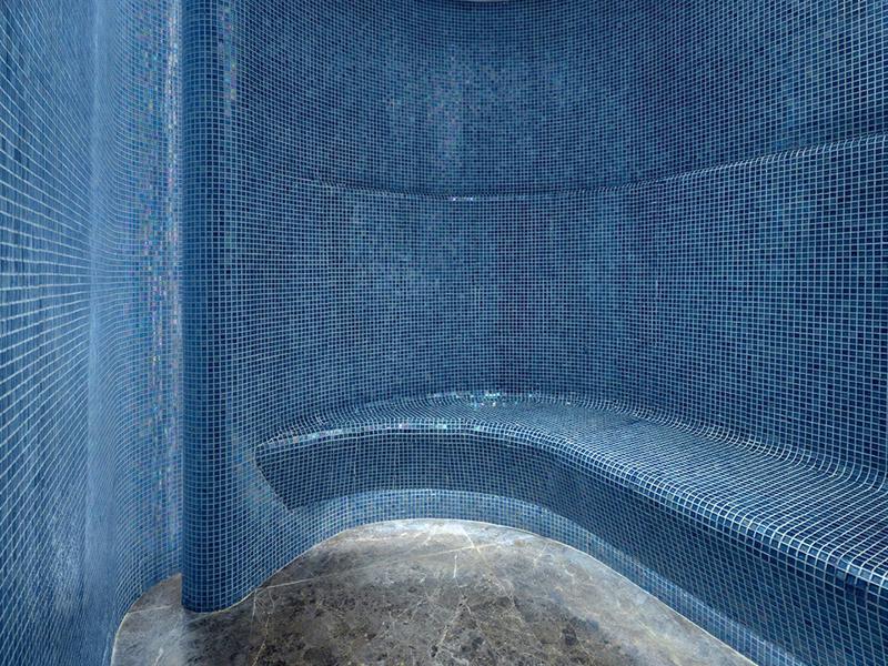 Parní místnost s modrými mozaikovými dlaždicemi a zakřivenou kamennou lavičkou.