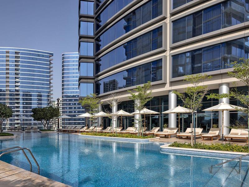 Moderní venkovní bazén s lehátky a slunečníky před skleněným mrakodrapem.